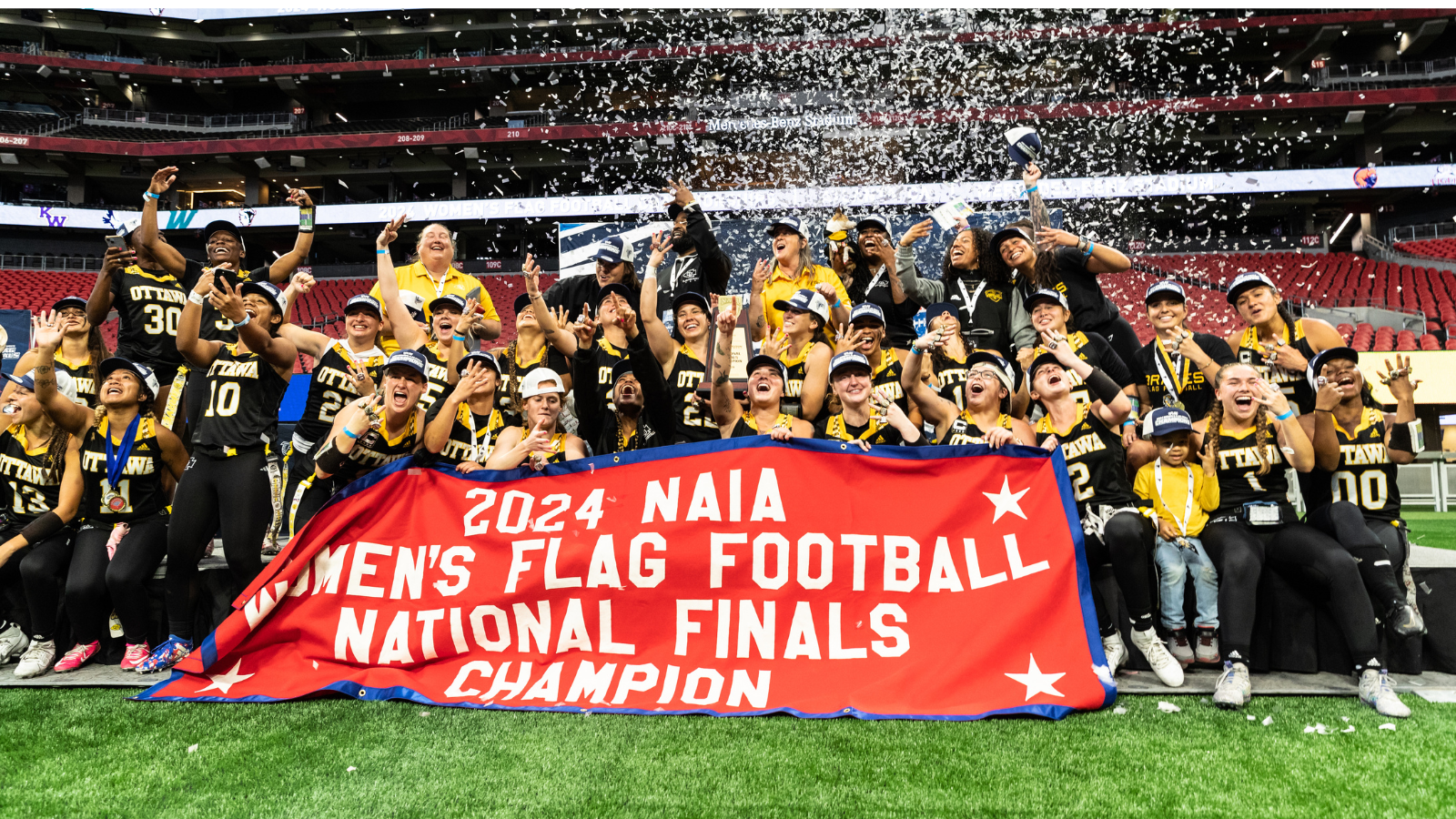 Ottawa University’s women’s flag football team celebrating in front of the 2024 NAIA National Championship red banner.